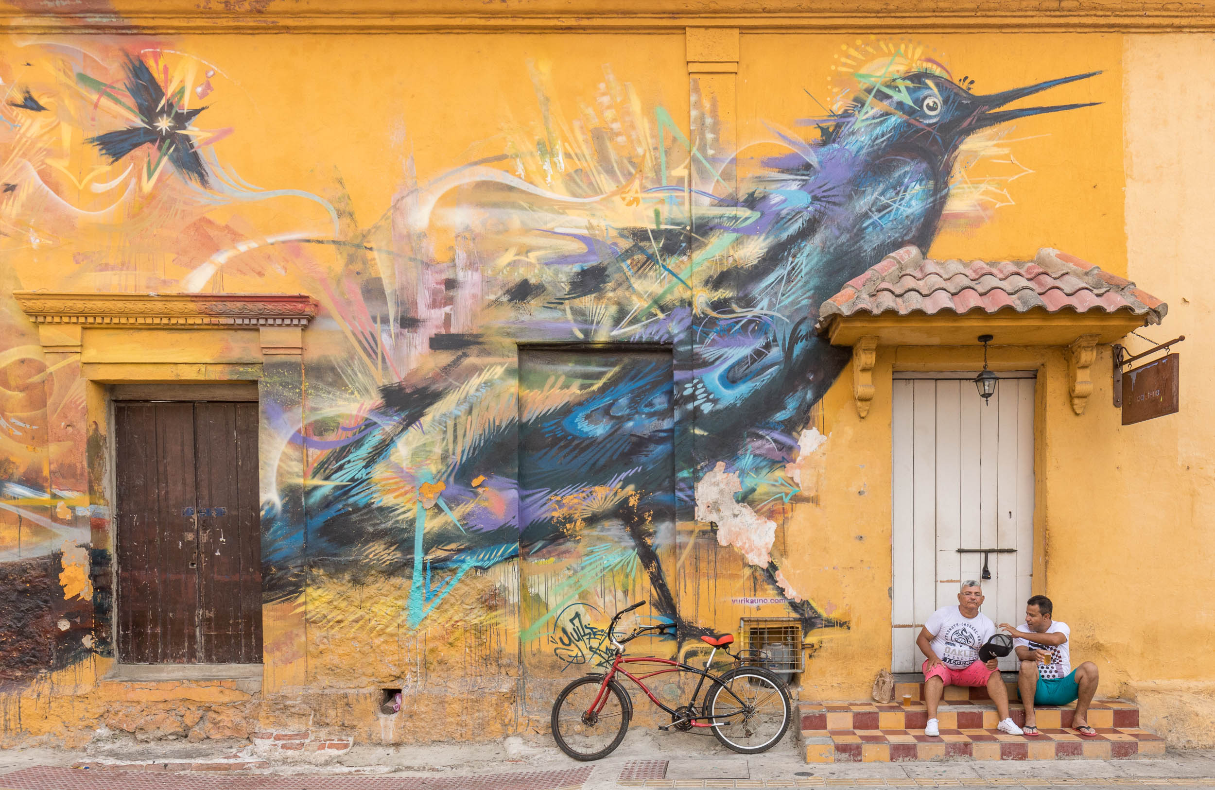Two men sitting in front of a huge mural of a bird.